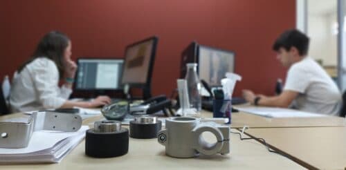 Dos técnicos trabajando en ordenadores en una oficina, con piezas mecánicas mecanizadas en primer plano sobre la mesa.