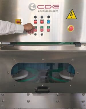 Operario con bata blanca accionando el panel de control de una máquina industrial de la marca CDE Equipos, con botones de colores y ventana de inspección.